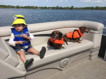 Image of a child wearing a hat and life jacket sitting on a bench in a boat with two dogs also wearing life jackets. Photo courtesy: Shannon Bauer   